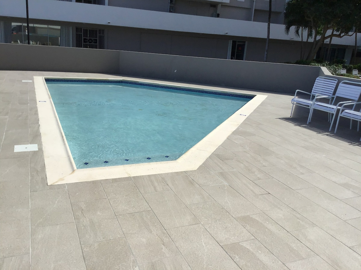 A clean, triangular swimming pool is set within a spacious tiled area. White lounge chairs are arranged nearby, providing a relaxing space for guests. The surrounding environment is bright and inviting, creating an open and airy atmosphere.