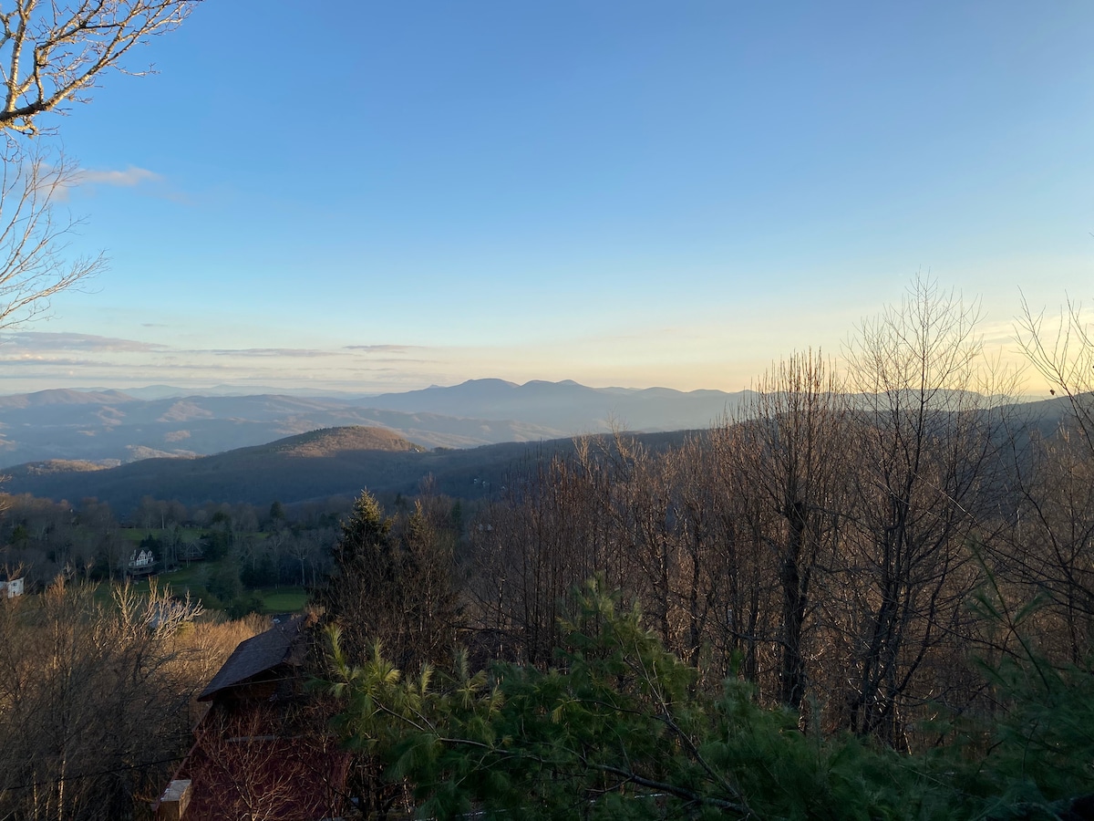 A serene view of mountain ranges is framed by bare trees, with a soft blue sky transitioning to warm tones at the horizon. The landscape features rolling hills and valleys, capturing the tranquility of the surrounding nature.