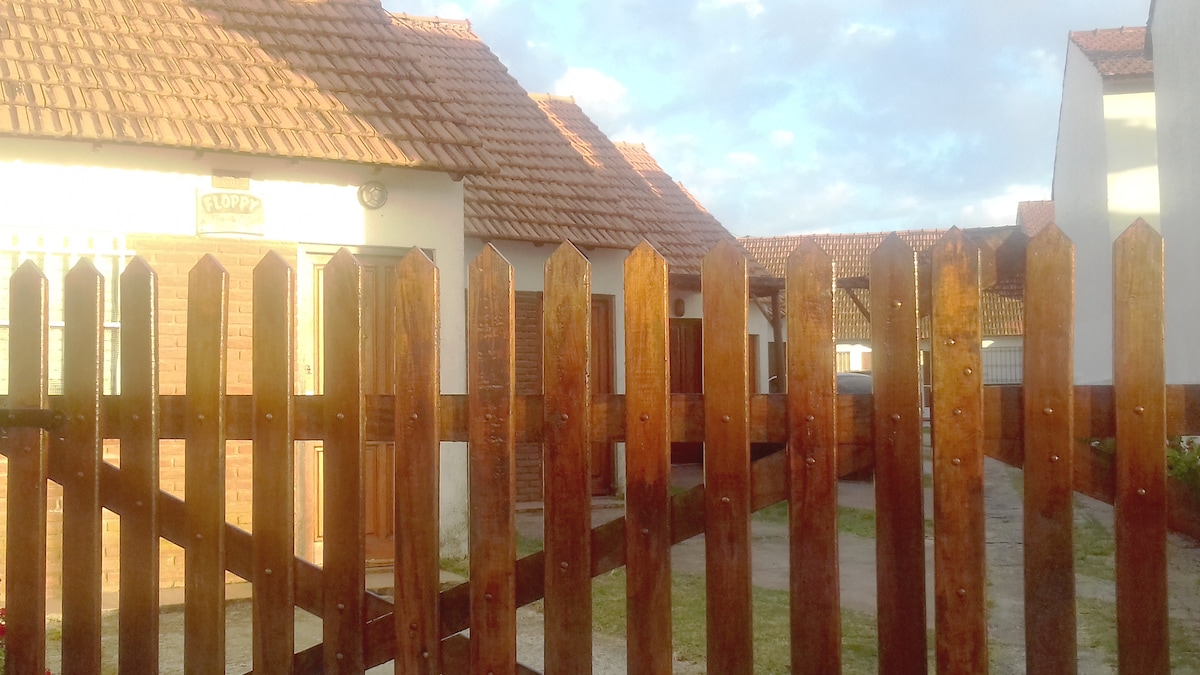 A wooden fence with pointed tops encloses a charming vacation home setting. The image depicts several rooftops with terracotta tiles under a partially cloudy sky. The entrance to the property features a sign, indicating the name, amidst a welcoming atmosphere.