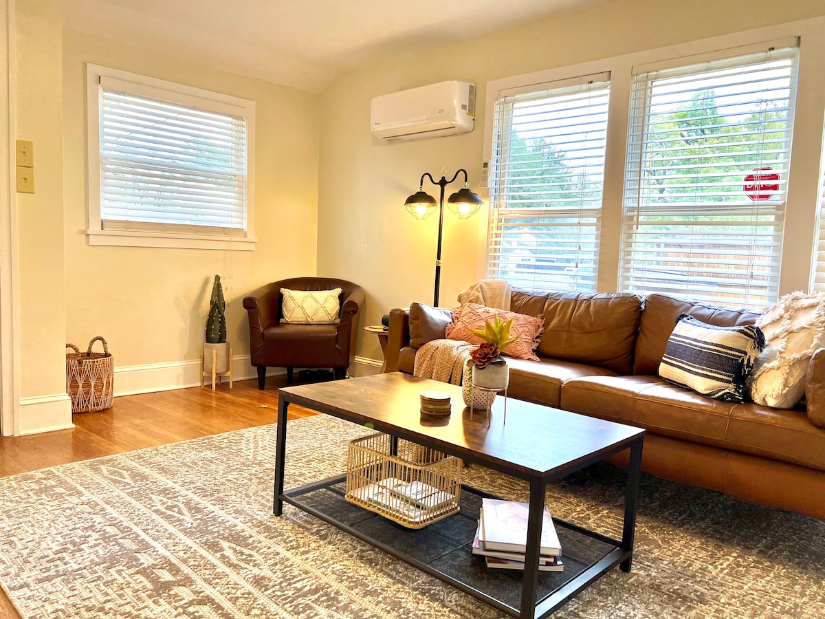 A welcoming living area features a brown leather sofa and an accent chair, positioned beside a coffee table with decorative items. Natural light enters through multiple windows with blinds, illuminating the hardwood floor and a patterned area rug.