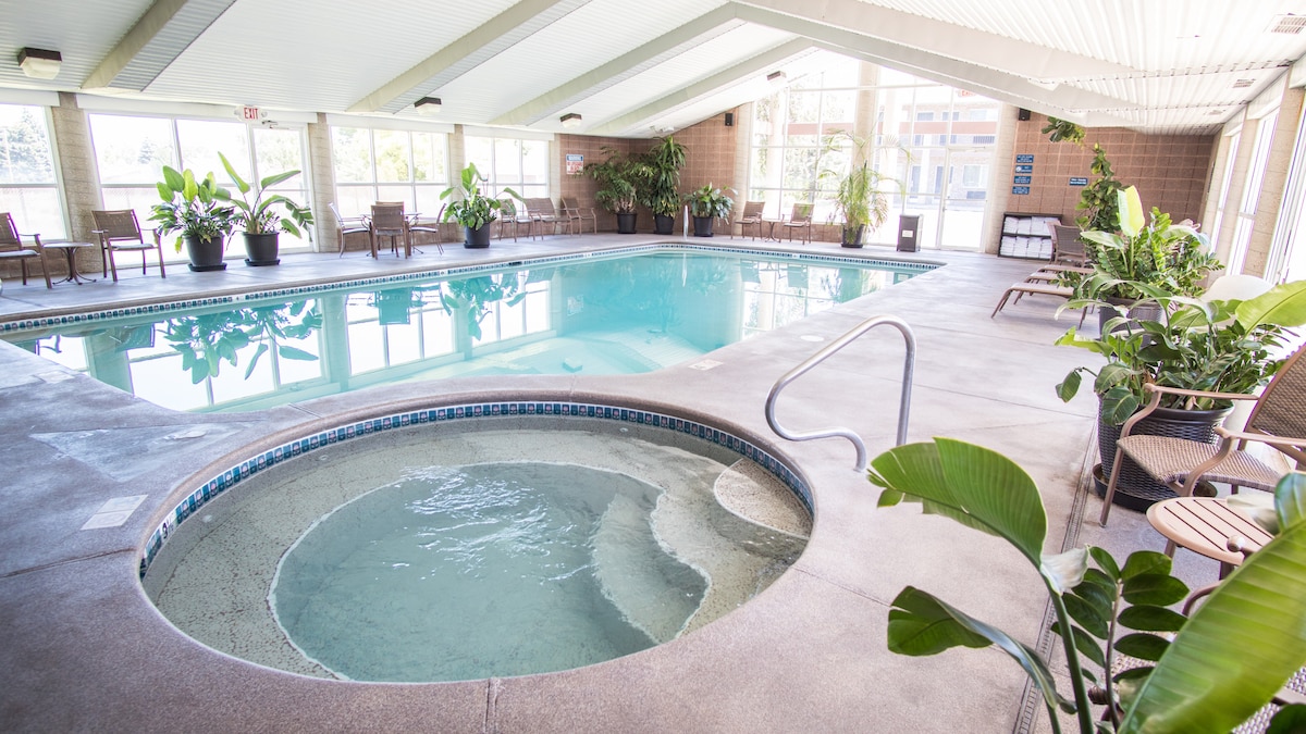 An indoor swimming pool features clear blue water, surrounded by large windows allowing natural light. A hot tub is positioned beside the pool, and several lounge chairs are placed throughout the area. Potted plants contribute to a refreshing atmosphere.