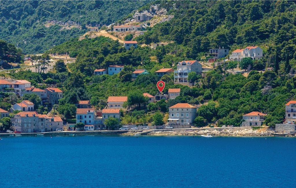 A coastal view showcases the serene bay of Sobra, lined with traditional buildings featuring terracotta roofs. The lush greenery of the surrounding hills complements the calm blue water, while the distant landscape adds depth to the tranquil setting.