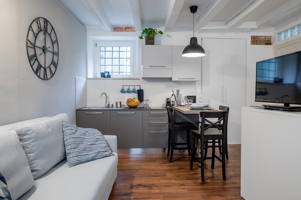 A modern kitchen area is paired with a small dining space, featuring a gray countertop and sleek cabinetry. A round black pendant light illuminates the space. The living area includes a light-colored sofa adorned with a decorative pillow, enhancing the cozy atmosphere.