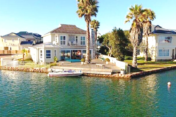 A serene villa is positioned along a canal, surrounded by neatly landscaped greenery and palm trees. A private dock holds a small boat, while large windows invite natural light into the spacious interior. The calm waters reflect the clear blue sky.