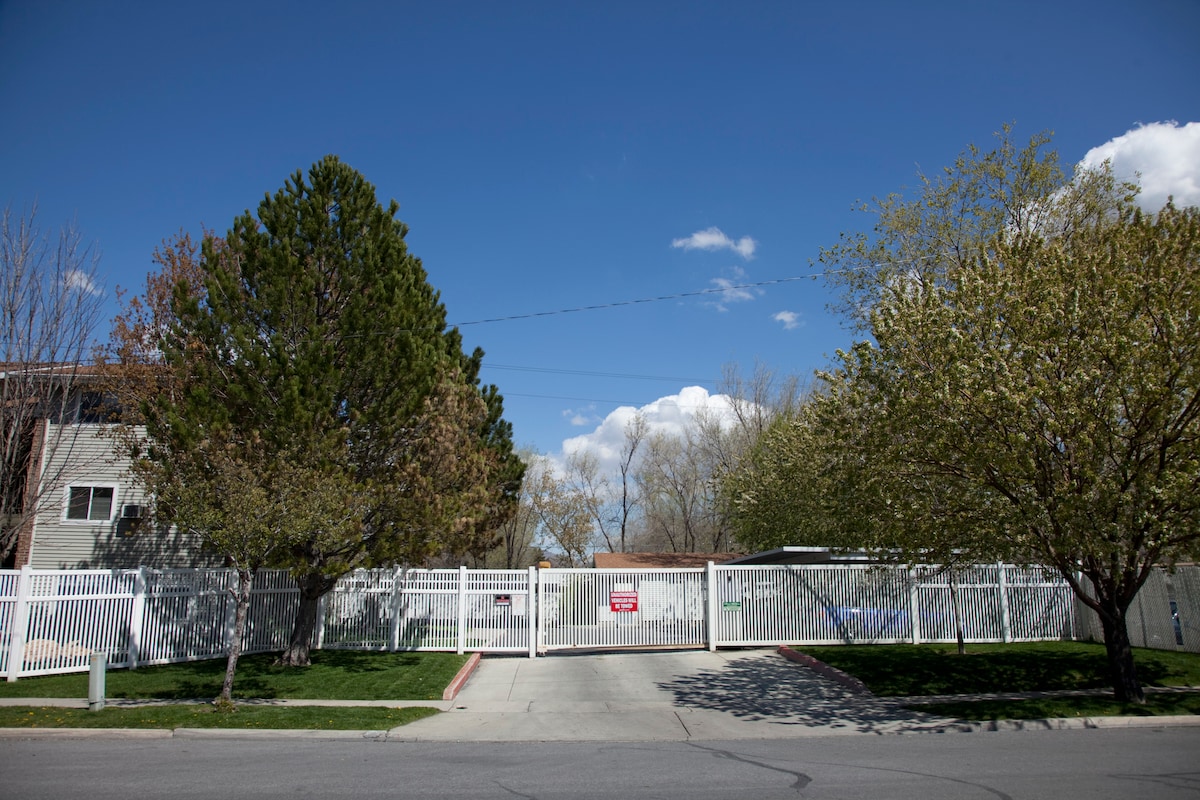 A gated entrance to the condo complex is visible, with a white fence enclosing a well-maintained landscape. Lush green grass and blooming trees provide a welcoming ambiance, complemented by a clear blue sky and fluffy white clouds in the background.