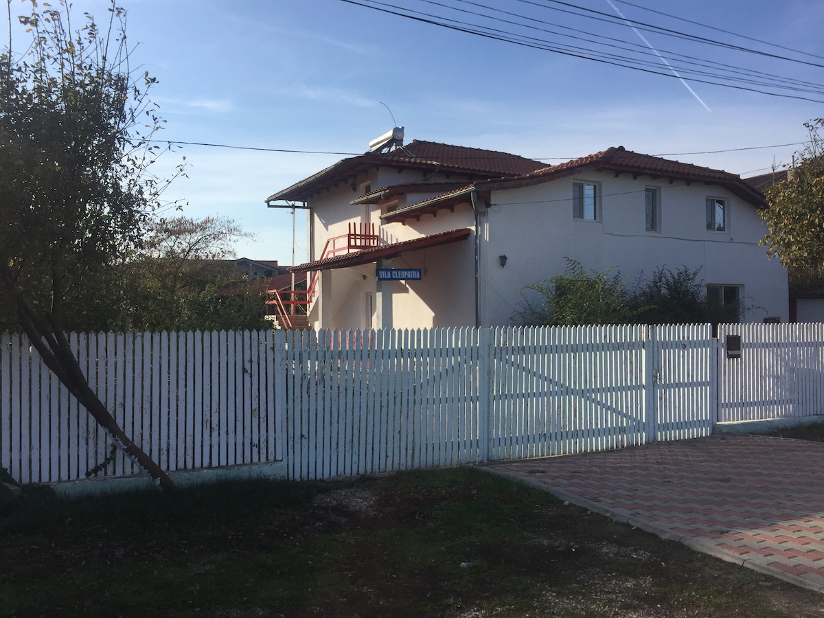A two-story house with a red roof is framed by a white picket fence. A set of stairs leads to a balcony on the upper level, while garden space is visible around the property. The clear sky offers natural light to the exterior.