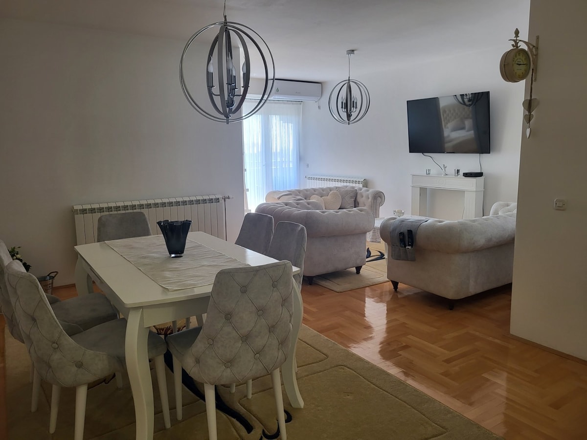 A light-filled living area features a comfortable seating arrangement with soft, neutral-toned furniture. A white dining table with upholstered chairs is positioned nearby. A wall-mounted television and decorative lighting fixtures add a modern touch to the space.