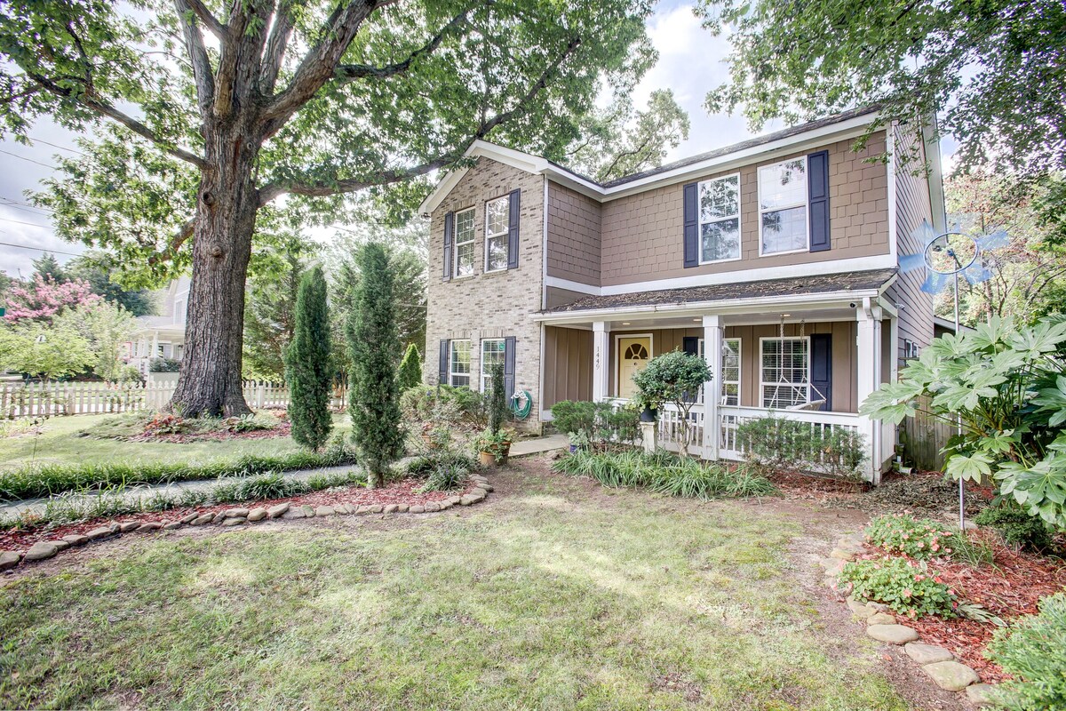 The exterior of a two-story home is framed by lush greenery and mature trees, including a large tree in the front yard. A neatly landscaped garden with stone edging surrounds the property, featuring manicured grass and decorative plants. A welcoming front porch is visible.