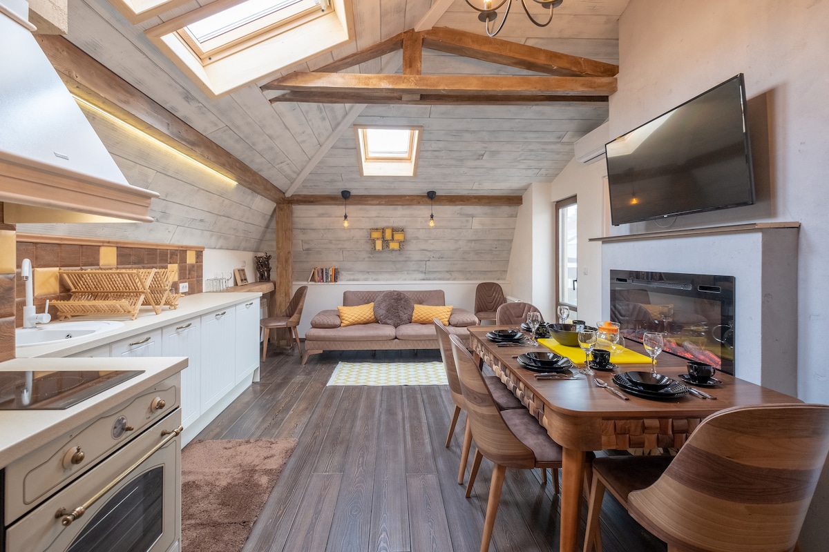 The spacious living area features sloped wooden beams and natural light from roof windows. A comfortable seating area includes a sofa, while a wooden dining table is set for meals. The open-plan kitchen is fully equipped with modern appliances, enhancing the inviting atmosphere.