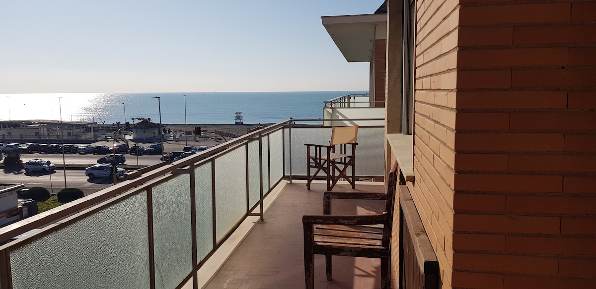 A spacious balcony overlooks the serene sea, featuring a wooden chair and a bench. The glass railing allows for unobstructed views, while the bright sunlight creates a warm ambiance throughout the outdoor area.