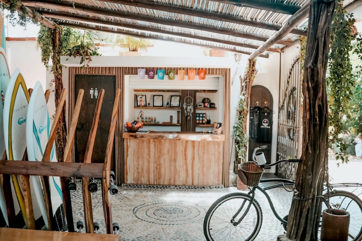 A welcoming reception area features a wooden counter, surrounded by surfboards displayed against the wall. Natural elements, such as a bicycle and greenery, are incorporated into the design, creating an inviting atmosphere for guests. Light filters through the open structure, providing a bright ambiance.