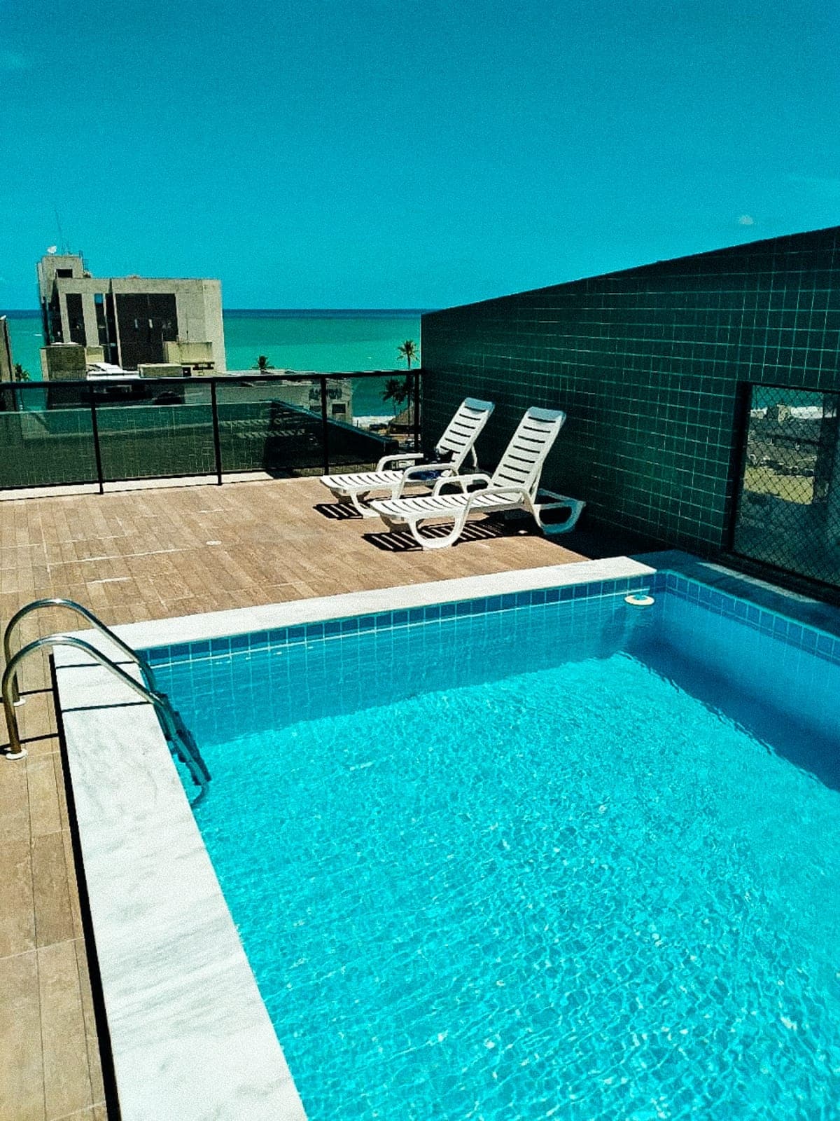 A rooftop area features a swimming pool surrounded by tiled walls and a wooden deck. Two lounge chairs are positioned near the pool, with a clear view of the ocean in the background, highlighting the inviting atmosphere of the space.