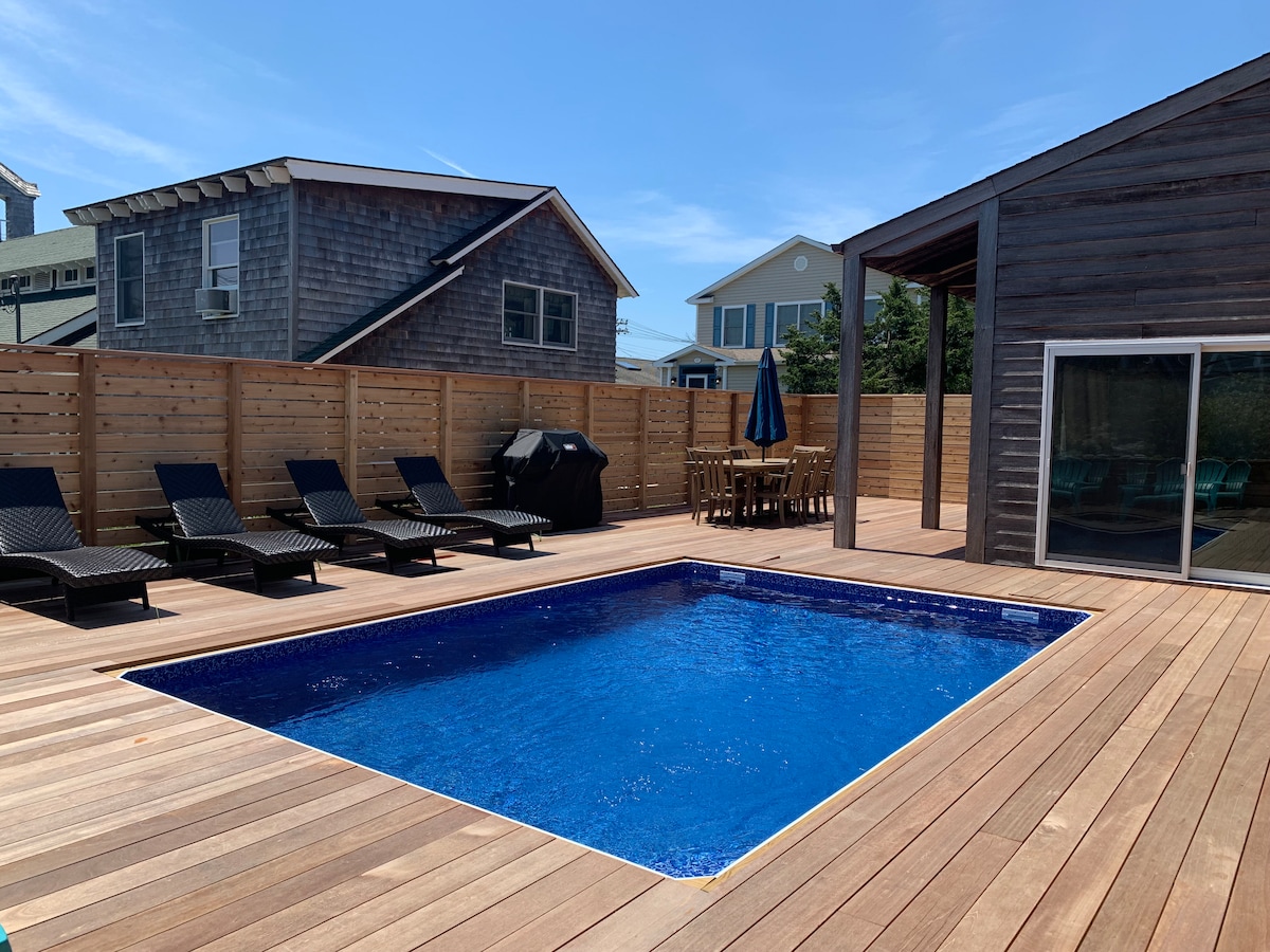 A newly constructed outdoor deck surrounds a heated salt-water pool, with five lounge chairs positioned on the wooden surface. A dining table with seating is visible nearby, and a large umbrella provides shade. The area is enclosed by wooden fencing, ensuring privacy.