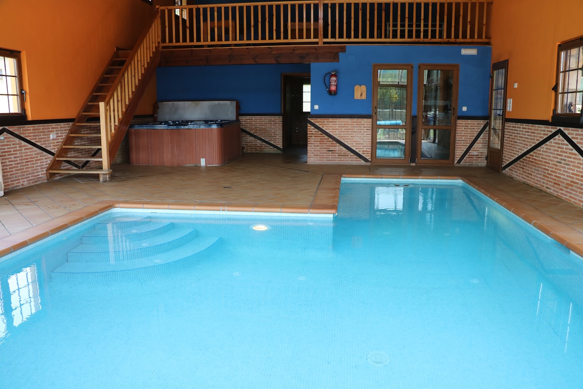 An indoor pool is surrounded by tiled flooring, with stairs leading to an upper level. Large sliding doors provide access to an outdoor area, while a wooden bar area is visible in the background. The walls are painted in vibrant orange and blue tones.