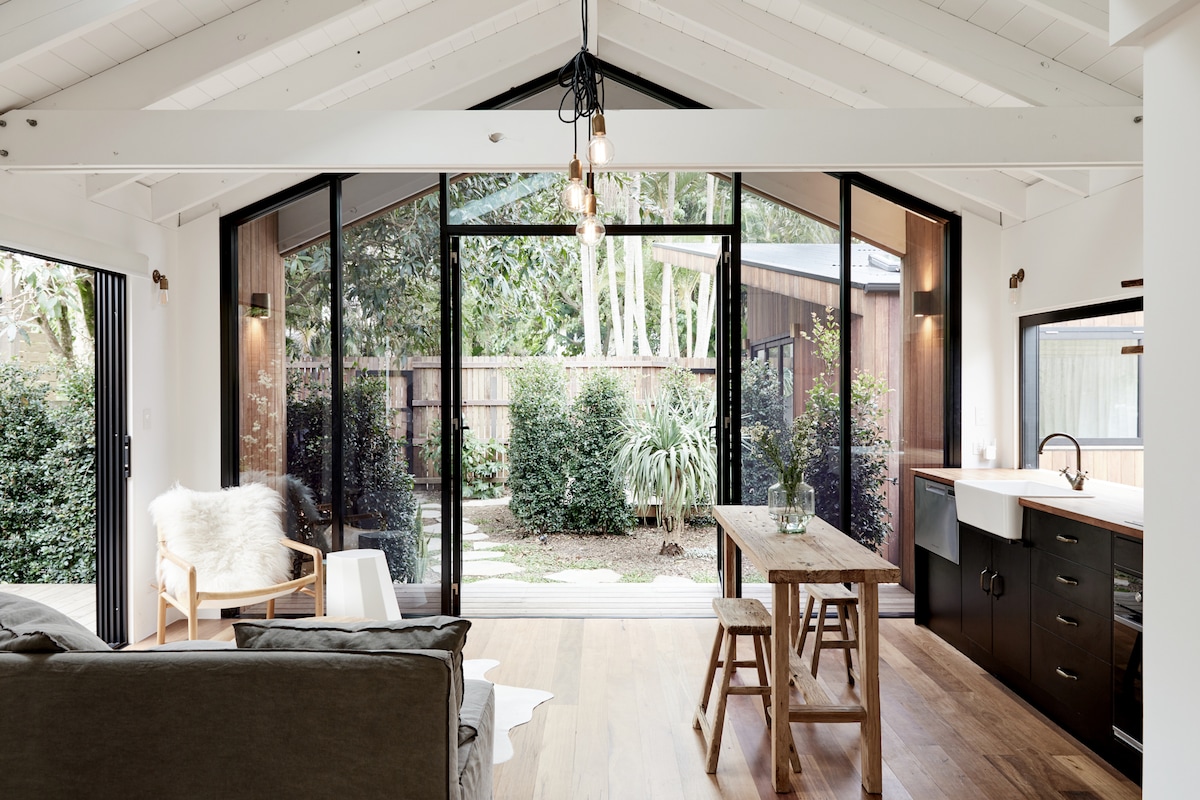 A studio interior features large, floor-to-ceiling glass doors that open to a serene outdoor space. Natural wood accents and rustic furnishings create a welcoming atmosphere. A spacious kitchen area, complete with a farmhouse sink, complements the dining space furnished with a wooden table and stools.