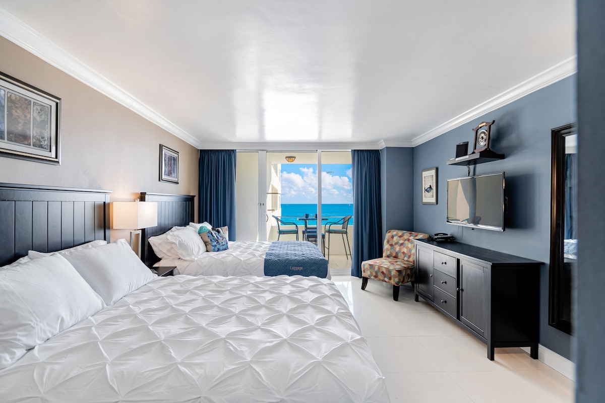 The studio features two queen beds dressed in white bedding, complemented by a modern blue accent wall. A flat-screen TV is mounted on the dark wood furniture, while sliding glass doors lead to a private balcony offering an ocean view. Natural light fills the space.