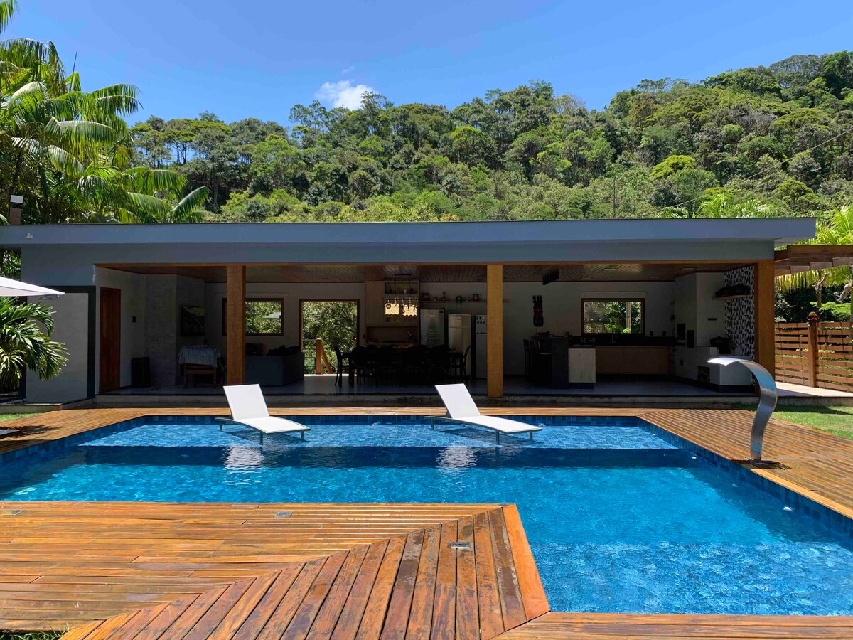 A serene outdoor area is showcased, featuring a swimming pool with two loungers positioned on wooden decking. The backdrop highlights lush greenery and mountains, while an open living space is visible through large windows. The setting promotes relaxation and connection with nature.