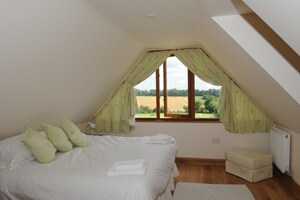 A spacious bedroom features a large bed adorned with soft pillows. Natural light filters through a triangular window, framed by green drapes, revealing views of the surrounding countryside. A small storage ottoman is positioned nearby, and light wood flooring adds warmth to the space.
