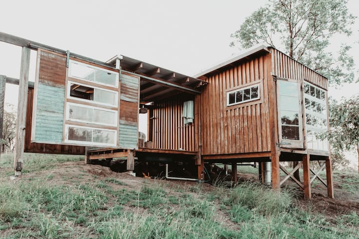 The image depicts a rustic cabin elevated on stilts with a unique design featuring a mix of wood and corrugated metal. Large windows bring in natural light, and the sliding doors add an industrial touch to the otherwise earthy, handcrafted structure. The cabin's elevated position and simple, open design blend harmoniously with its natural surroundings, creating a peaceful retreat that feels both modern and rustic. It appears to be set in a rural, grassy area, offering a perfect escape into nature.