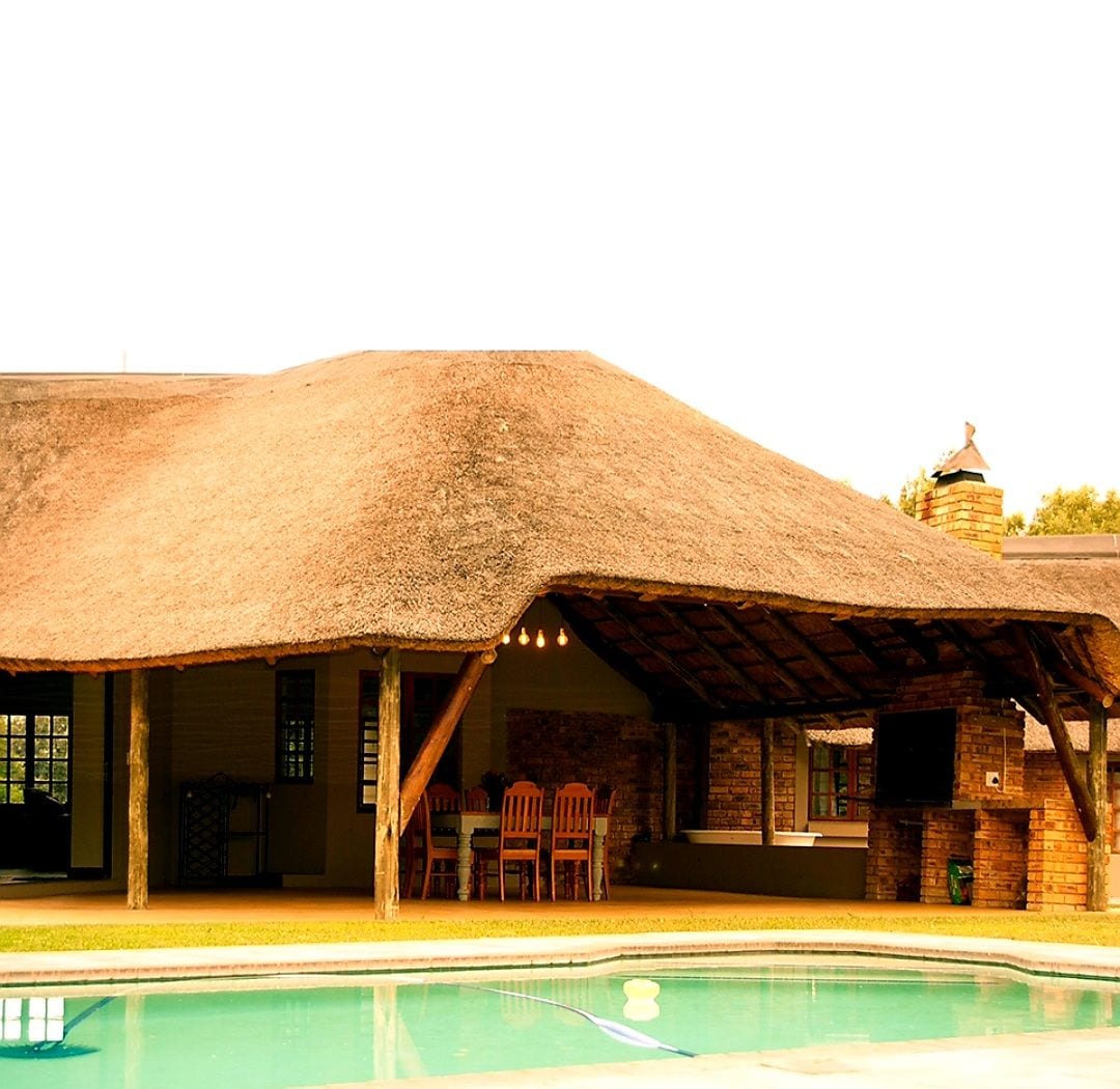 An expansive outdoor entertainment area features a thatched roof supported by wooden beams, providing shade for a dining table and chairs. A swimming pool reflects the structure and surrounding landscape, enhancing the outdoor atmosphere.