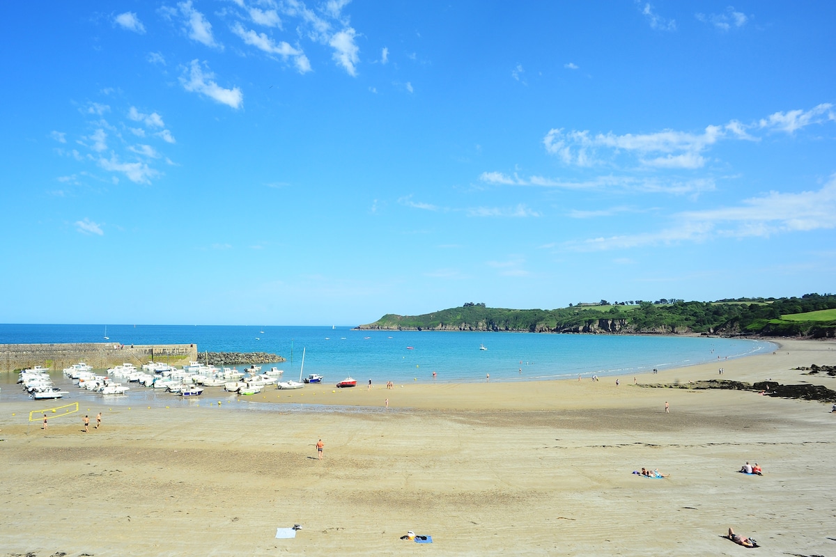 The image captures a sandy beach lined with calm turquoise waters, featuring a small harbor with boats. Lush green hills rise in the background under a clear blue sky, providing a serene coastal landscape. Sunbathers are seen enjoying the warm sun along the shoreline.