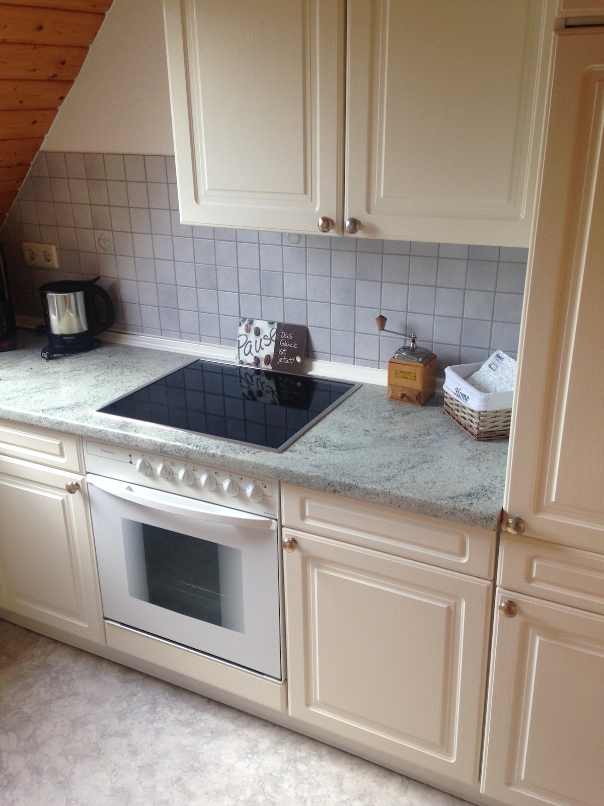 A functional kitchen space features light cabinetry and a grey tiled backsplash. An electric stovetop and an oven are installed in the countertop, alongside a kettle. A small basket and paper items are placed on the counter, enhancing the organized appearance.