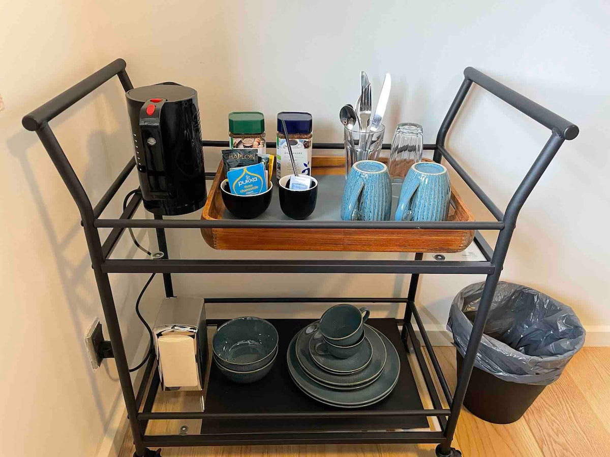 A functional cart is visible, featuring a tray with various glassware, utensils, and a kettle. Below, a selection of dishes and bowls is neatly arranged, while a trash bin is positioned alongside the cart. The setup promotes convenience for guests.