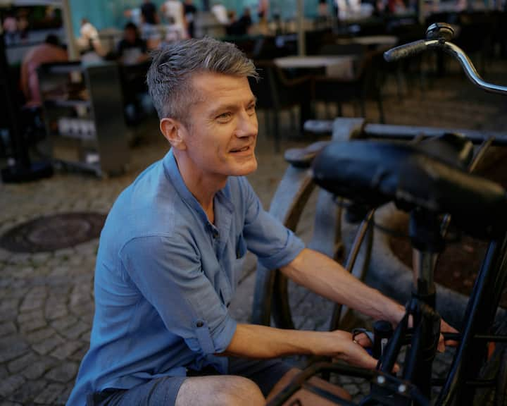 Un hombre con camisa azul de rodillas frente a una bicicleta intenta abrir un candado en forma de U.