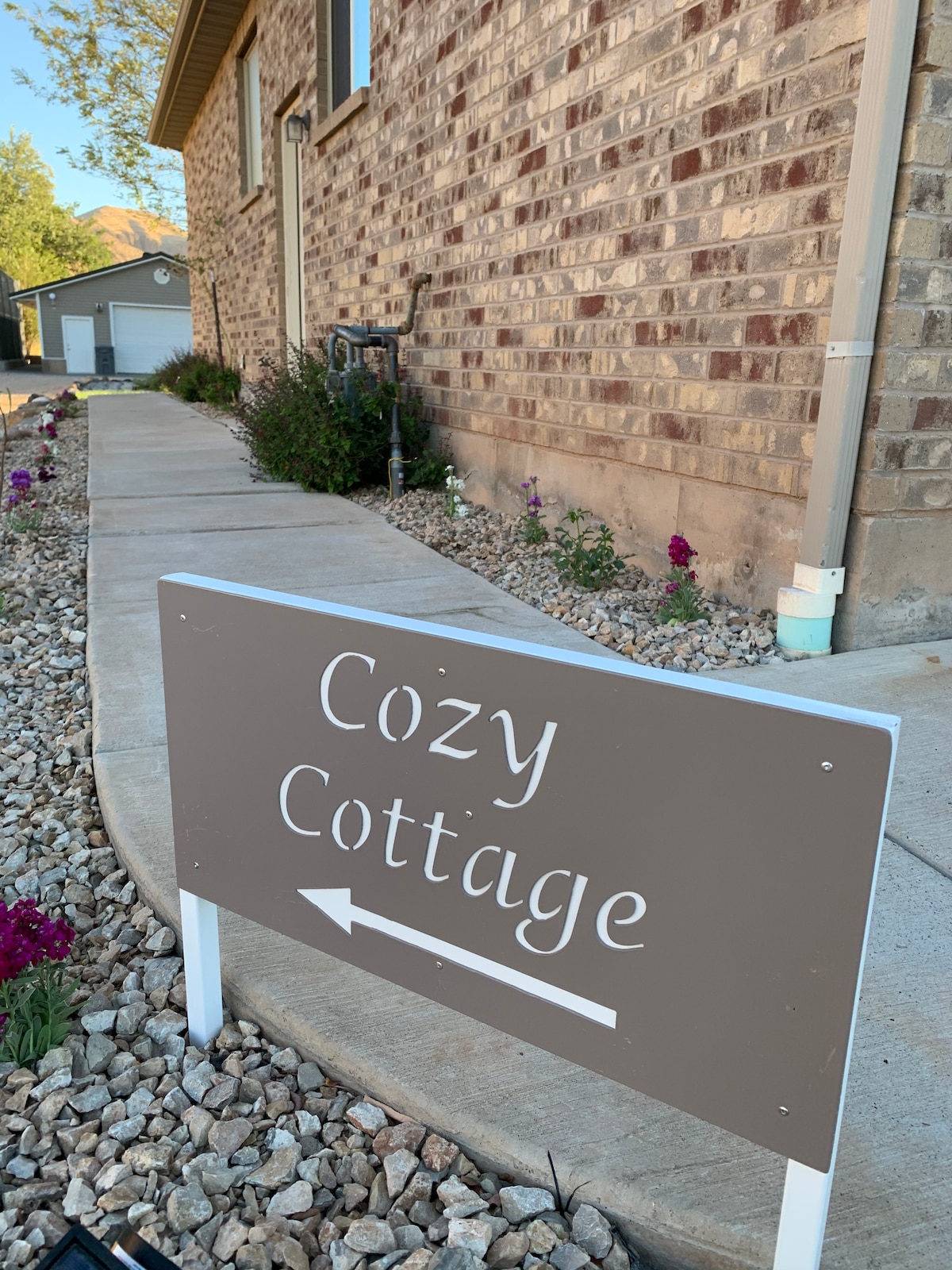 A welcoming sign is situated at the edge of a well-maintained pathway, indicating the direction to the Cozy Cottage. Colorful flowers are seen along the side, contributing to the inviting appearance of the walkway, which is lined with gravel.