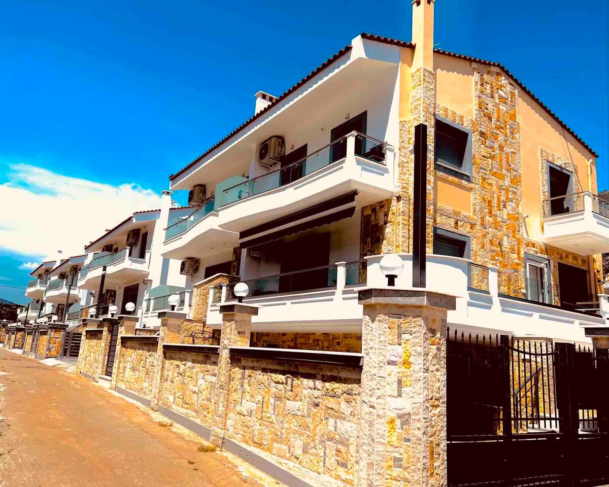 A contemporary building features multiple balconies with glass railings, showcasing a blend of light-colored stucco and stone accents. The structure is situated along a paved pathway, and air conditioning units are visible on exterior walls.