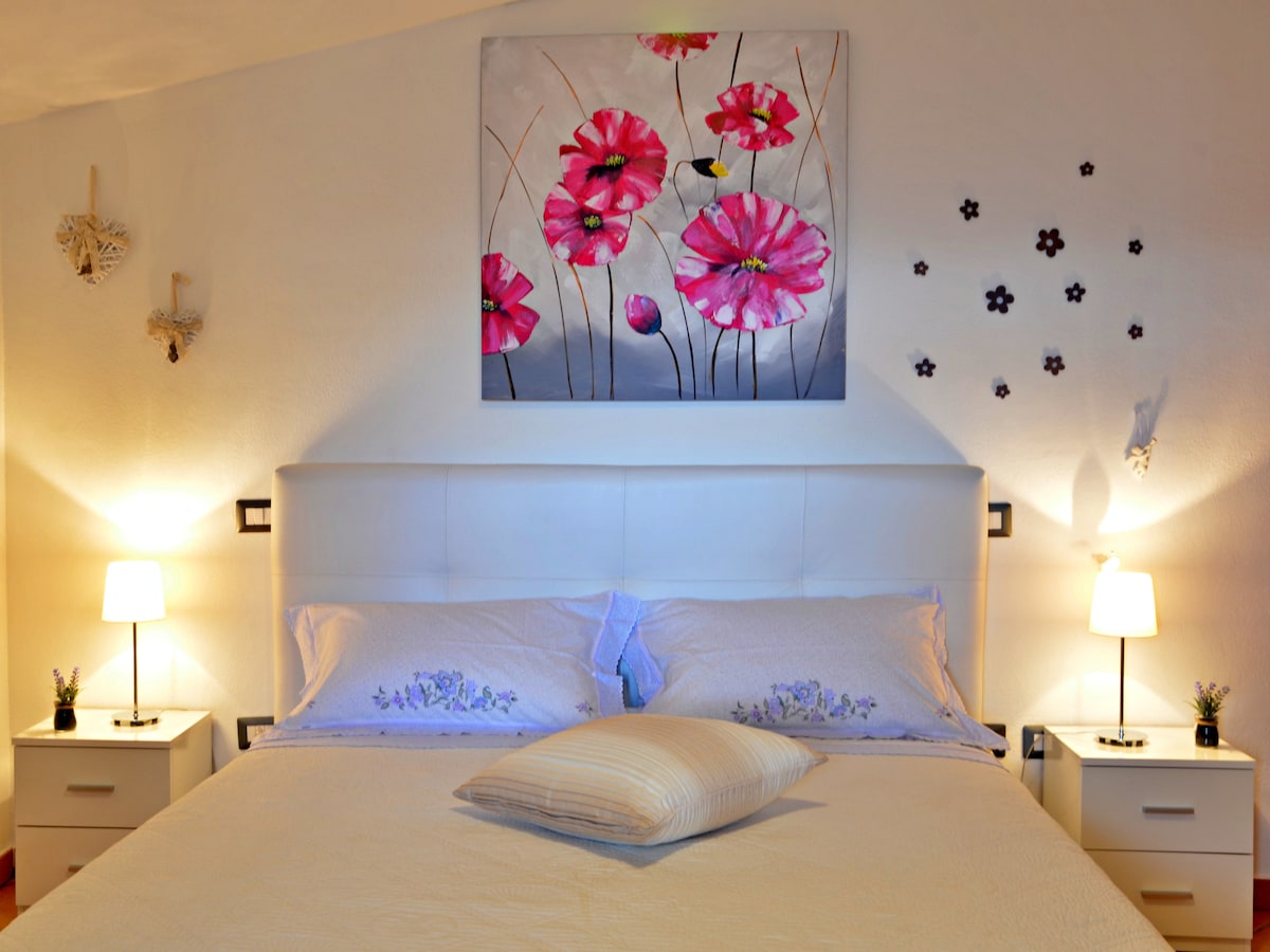A spacious bed is positioned against a white wall, complemented by bedside tables with lamps on each side. Above the bed, a floral painting in vibrant colors attracts attention, while smaller decorative elements are arranged on the wall, adding charm.