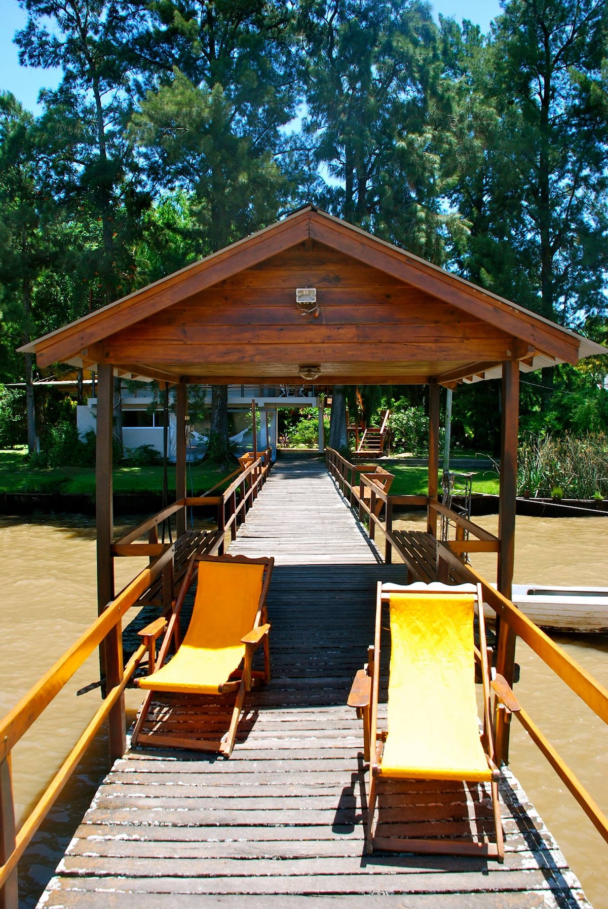 A wooden dock extends over the water, featuring two yellow lounge chairs positioned for relaxation. Surrounded by lush greenery, the structure leads towards a covered area, offering a serene view of the river and inviting a peaceful atmosphere.