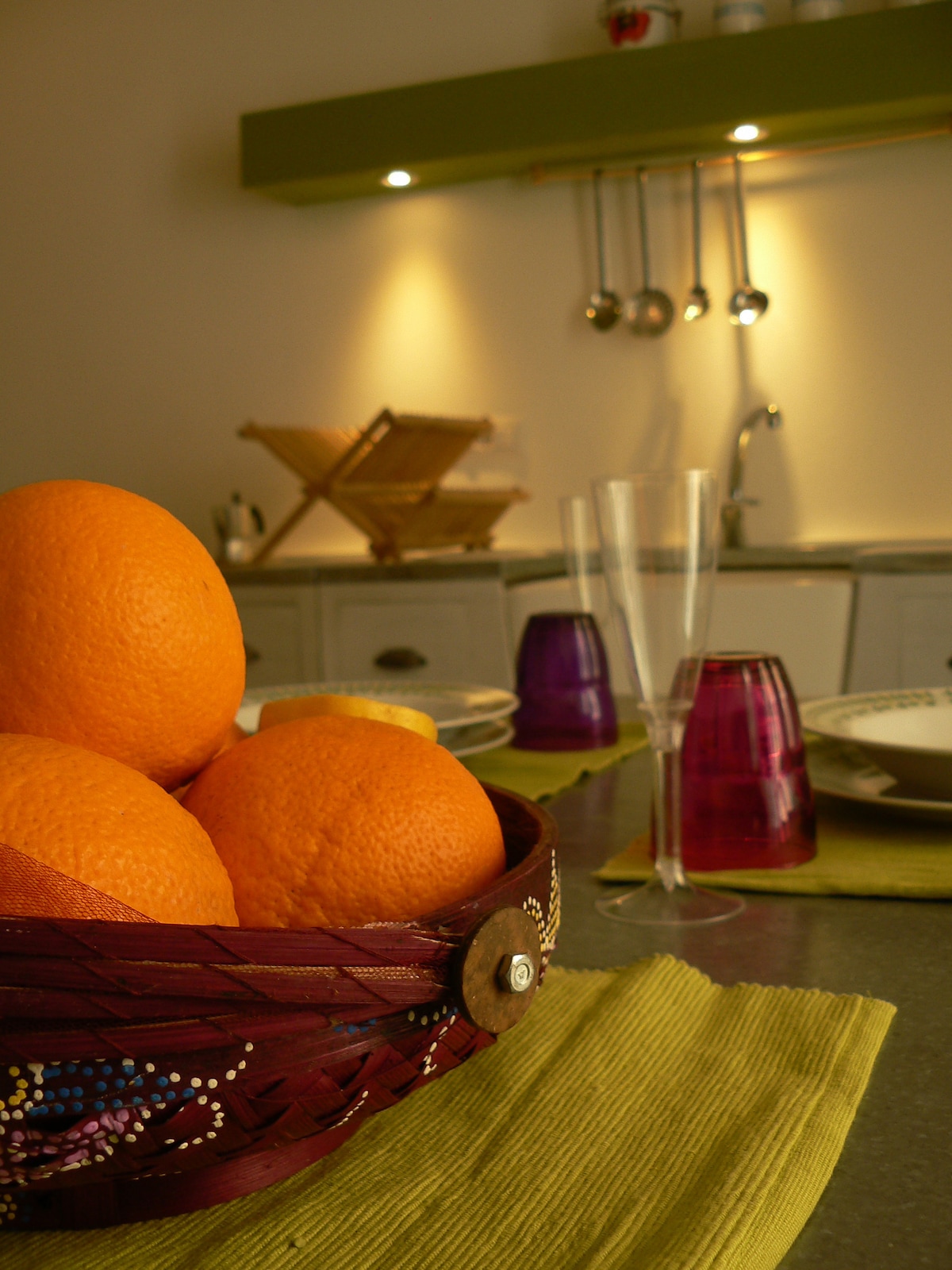 A woven basket holds several oranges, positioned at the forefront. A subtle green counter provides a backdrop, while a wooden drying rack and kitchen utensils are displayed in the background. Glassware and plates are set on textured green placemats, enhancing the kitchen's functional charm.