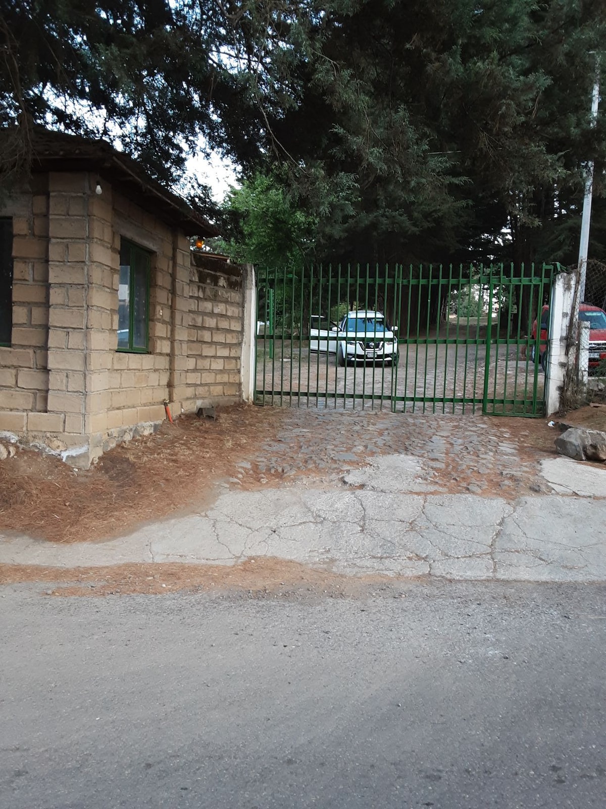 An entryway is framed by a green metal gate, offering a view of a gravel pathway leading to parked vehicles. The stone wall of a nearby structure is partially visible on the left, with trees providing a natural border on the property.