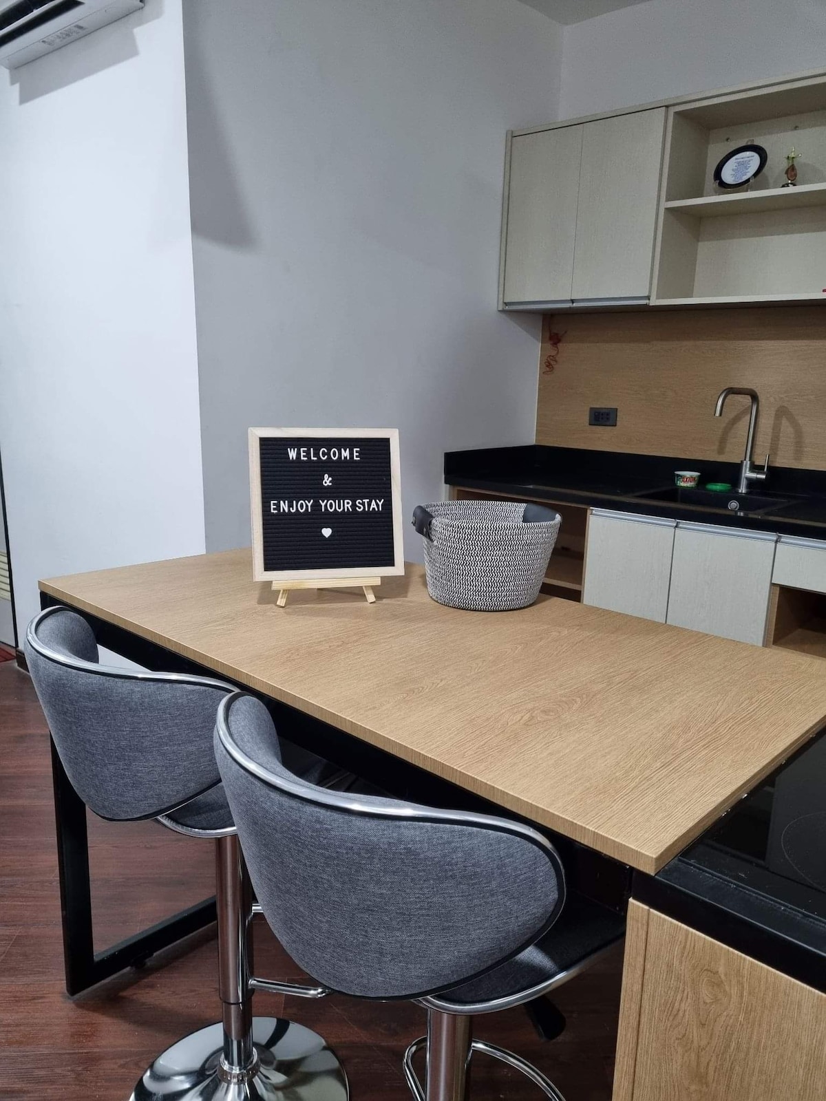 A modern kitchen area is showcased with an island-style table and two bar stools. A welcome sign is placed on an easel, and a woven basket is positioned on the tabletop. The kitchen counters feature a sleek black design, with a sink and simple cabinetry visible.