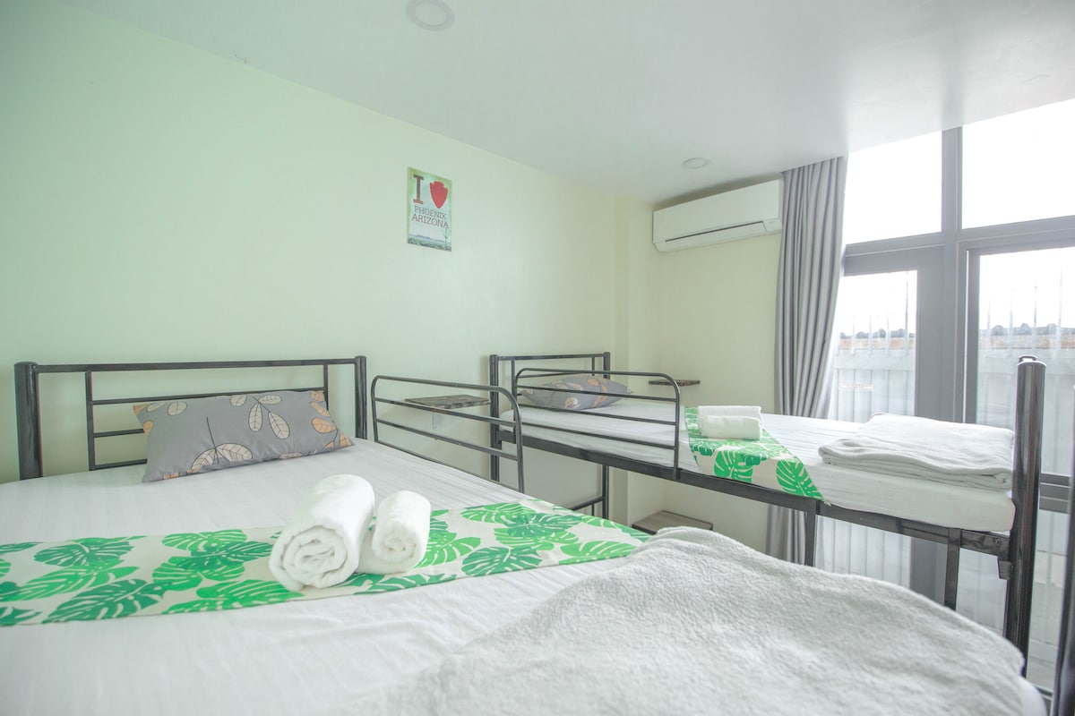 A bright room features a spacious bed and two additional beds, each with simple metal frames. Natural light fills the space through large windows adorned with light curtains. Soft towels and a decorative blanket with leaf patterns are neatly arranged on the beds.