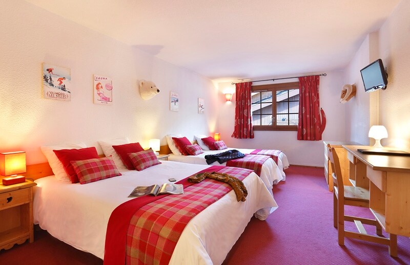 A well-appointed family room showcases three beds, featuring crisp white linens and red checkered accents. A wooden desk and chair provide a workspace. Natural light filters through a window adorned with patterned curtains, illuminating a warm atmosphere. Decorative wall art adds character to the room.