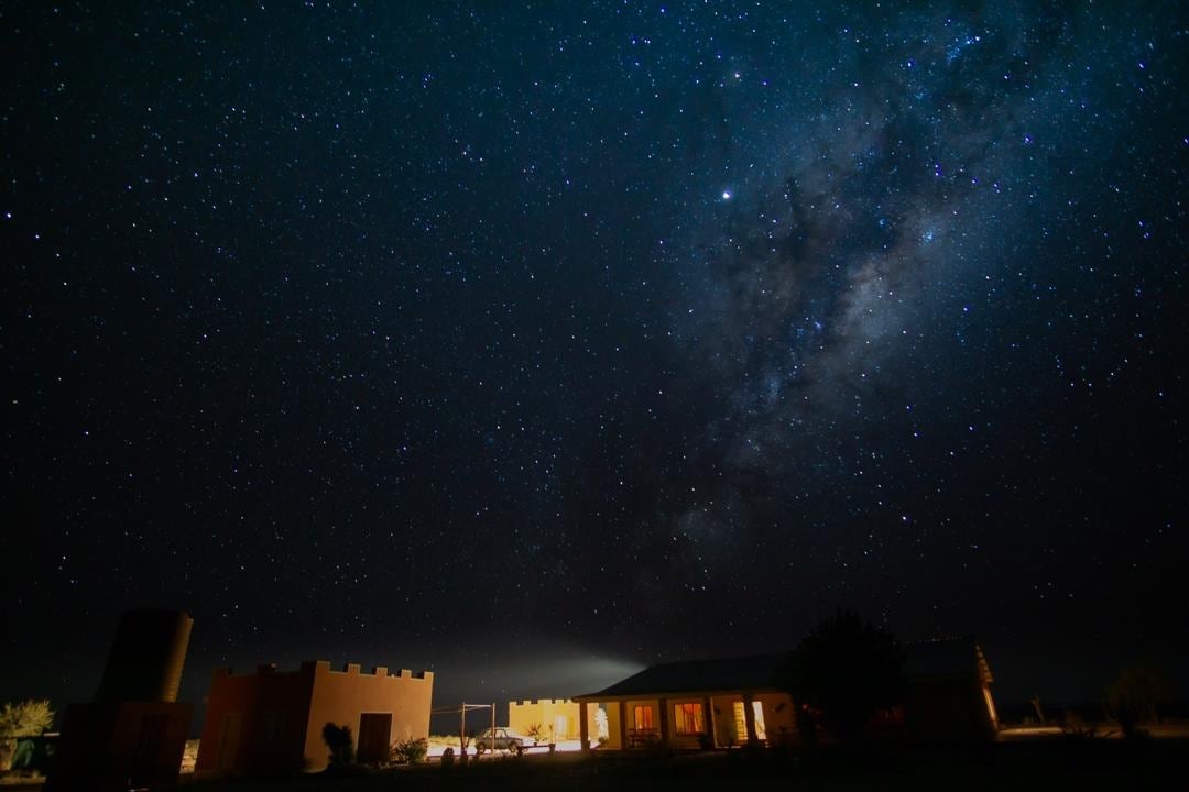 A night sky filled with stars and the Milky Way is visible above a tranquil building. The structure features warm-colored walls and illuminated windows, set against the dark silhouette of the landscape.