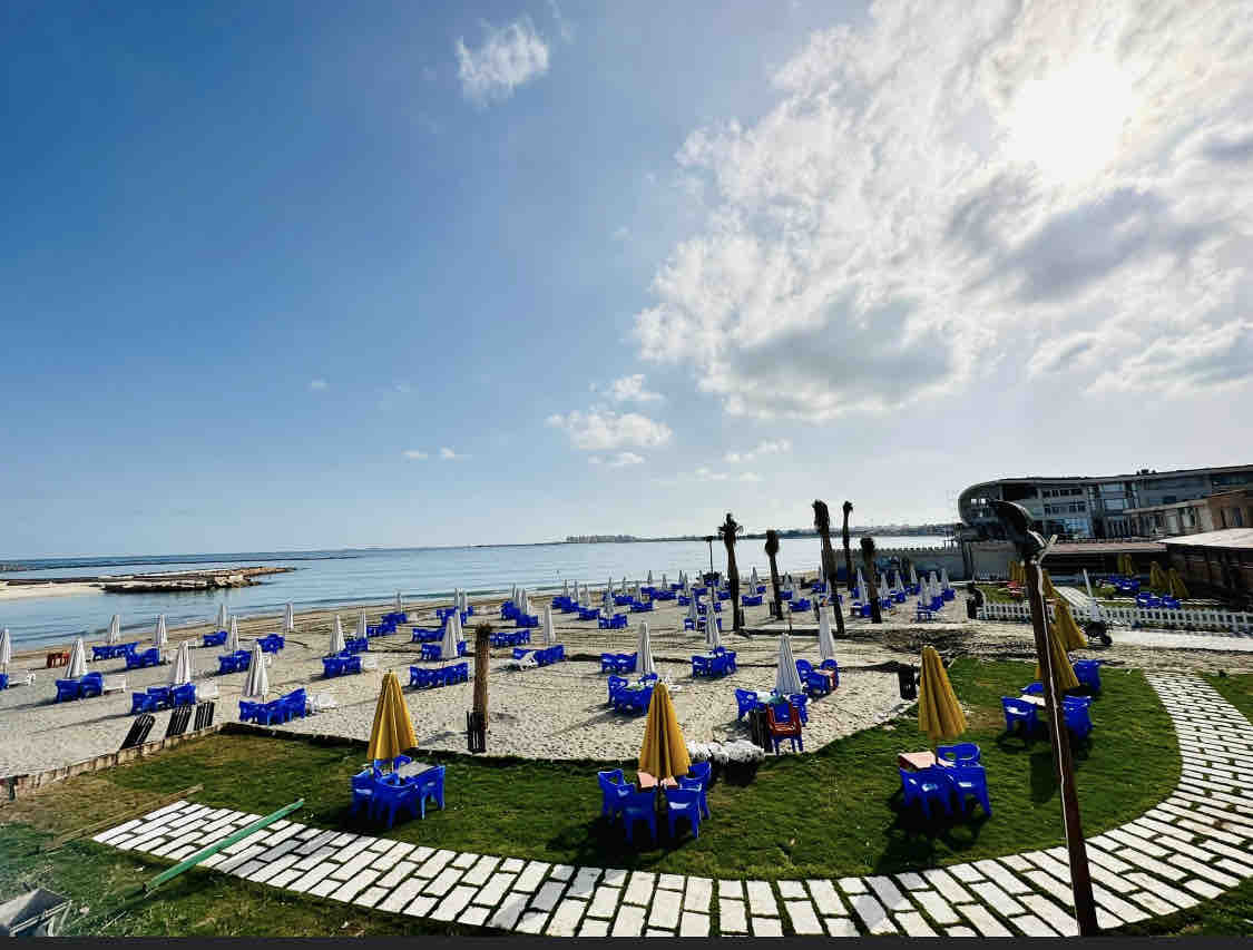 A sunlit beach area features numerous blue lounge chairs arranged under yellow umbrellas. A pathway of stone pavers leads through the grassy space towards the water, with the sea visible extending into the distance under a clear sky.