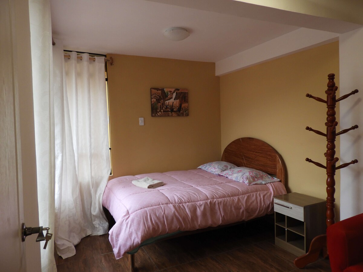 A cozy room features a double bed with a light pink blanket, complemented by floral bedding. Natural light filters through a sheer curtain, illuminating the space. A wooden nightstand sits beside the bed, and a coat rack is positioned against the wall.