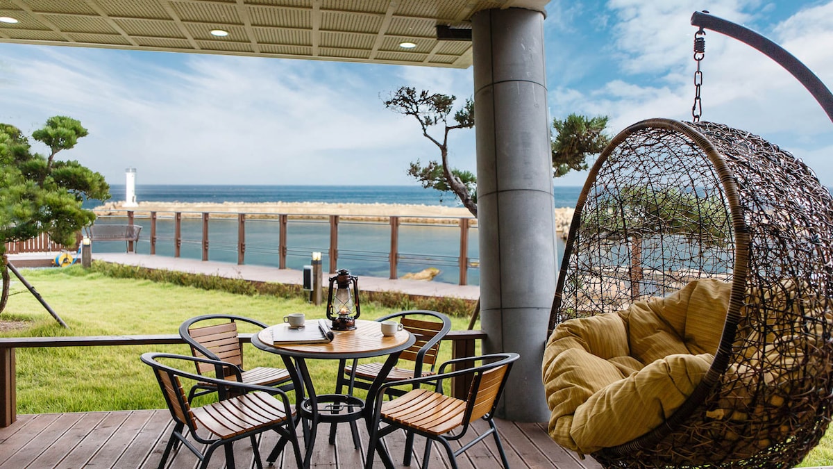 A cozy outdoor seating area features a round hanging chair and a small table surrounded by four wooden chairs. The scenic view displays a calm body of water with a distant lighthouse and a well-maintained grassy area, perfect for relaxation.