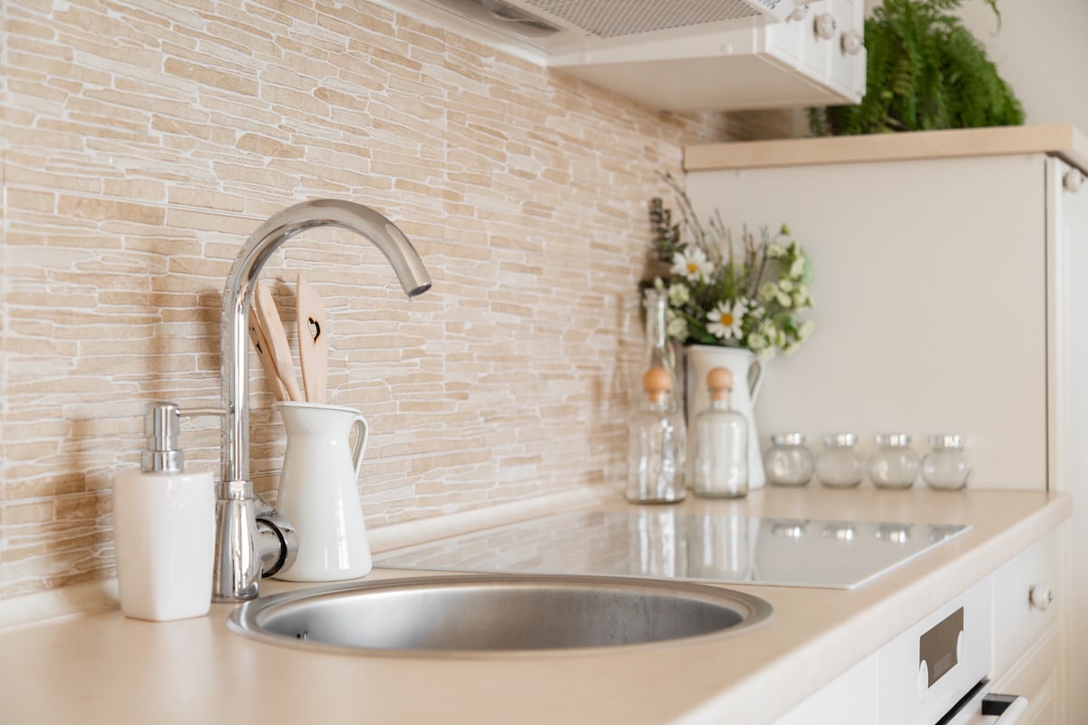 The kitchen features a circular stainless steel sink accompanied by a sleek faucet. Cooking utensils are neatly arranged in a container, while decorative glass jars and a small vase of flowers add a touch of elegance. Natural stone tiles create a warm backdrop.