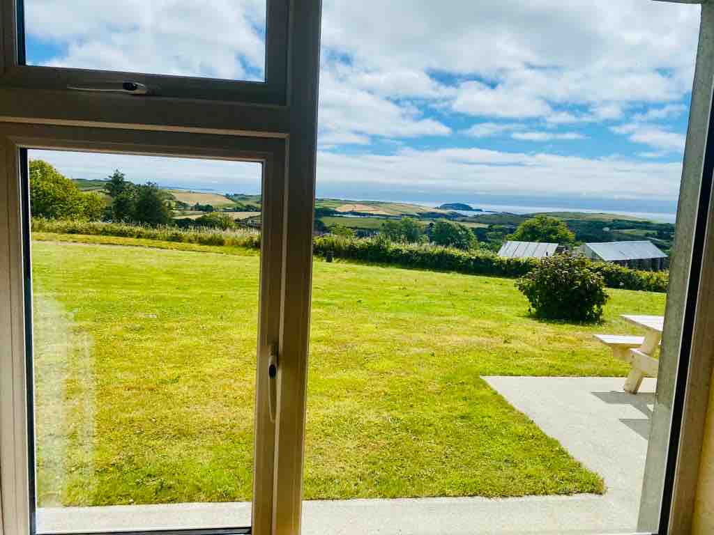 A view from an interior window showcases a lush green lawn that leads to scenic hills and a distant coastline. Soft clouds drift across a bright blue sky, enhancing the serene outdoor landscape.