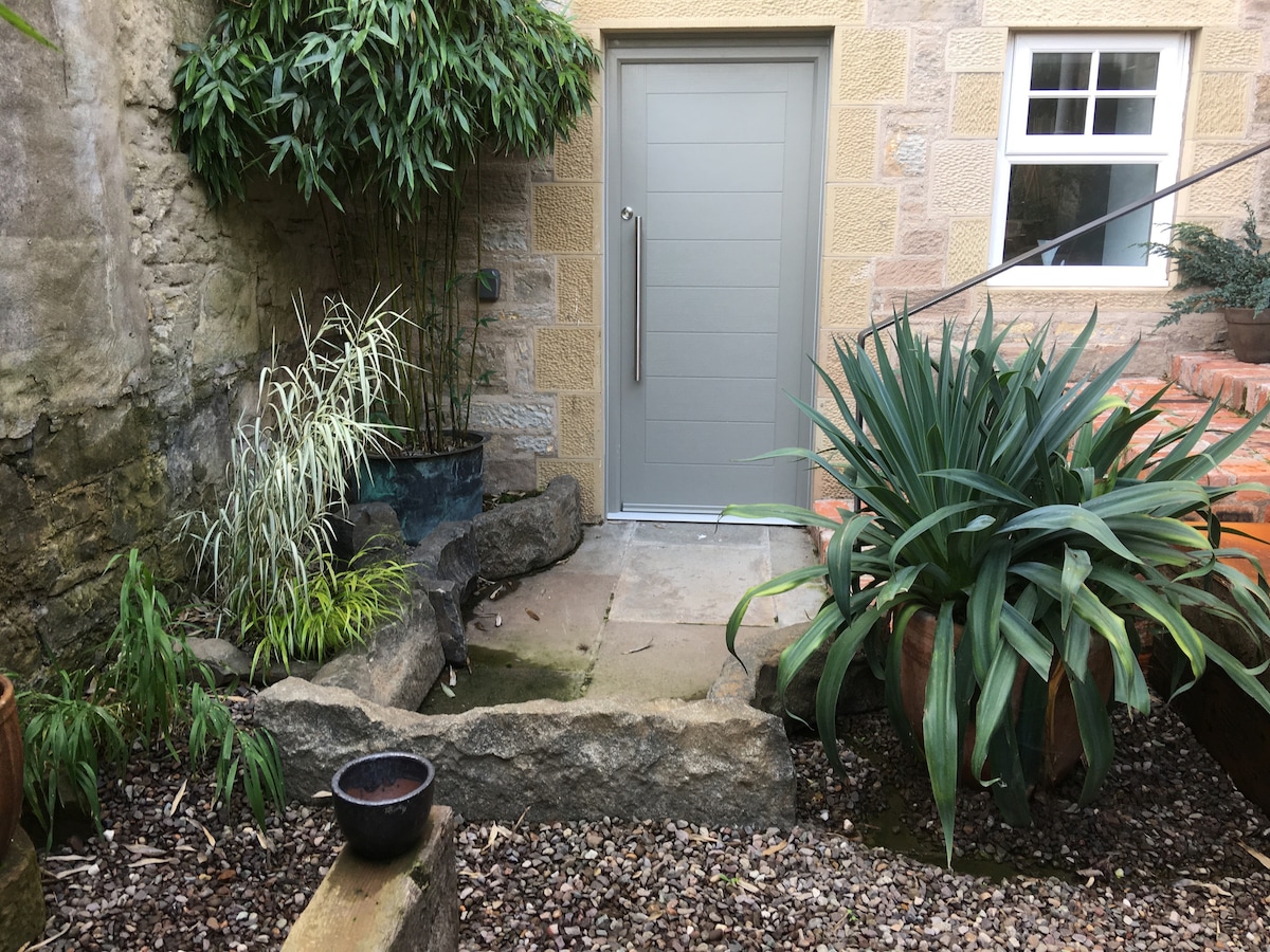 A stone pathway leads to a contemporary grey door, framed by an assortment of greenery, including tall plants and bamboo. Smaller pots with lush foliage are positioned around the area, creating a serene and vibrant garden space.