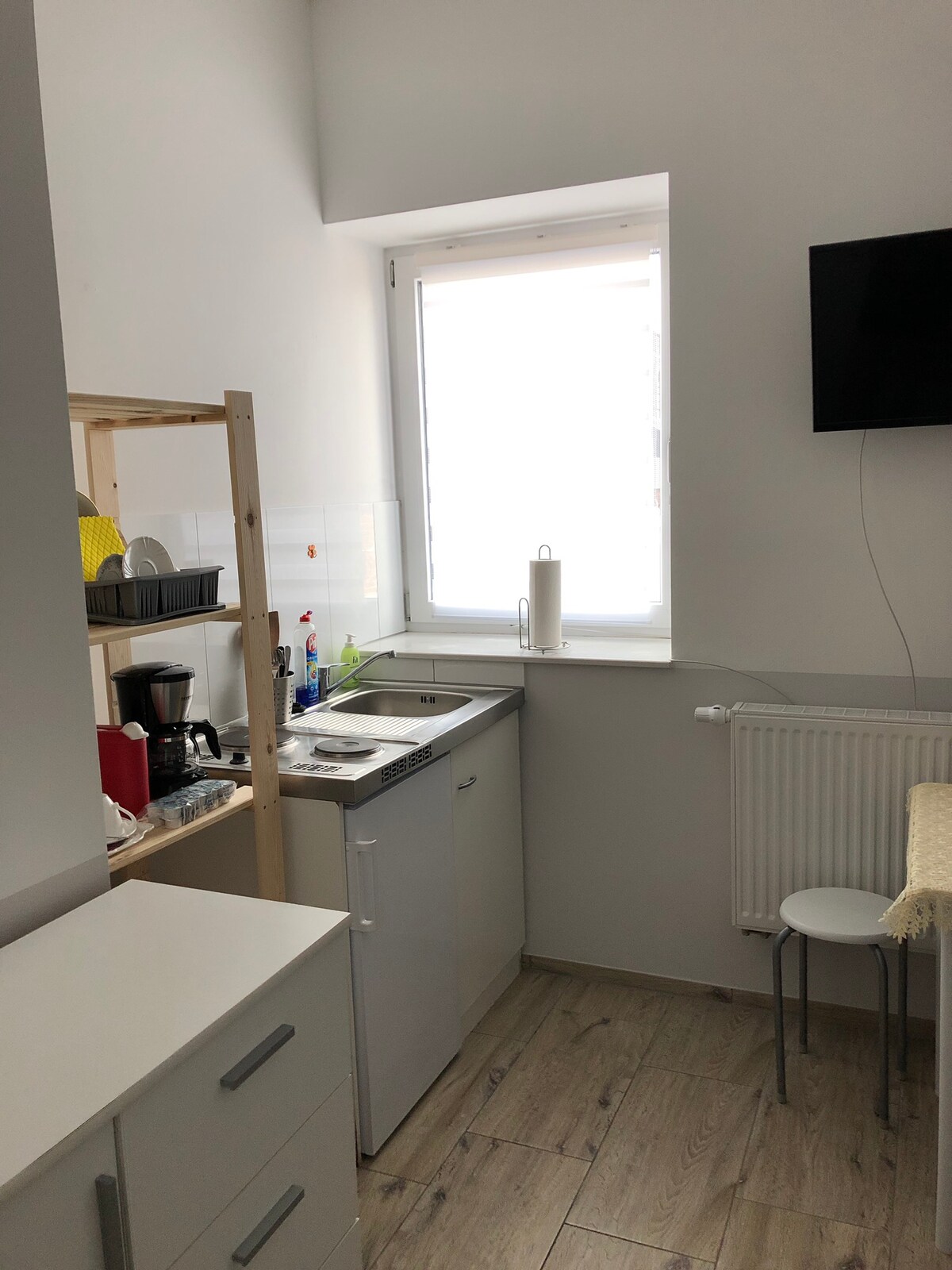 A compact kitchenette is equipped with essential appliances, including a coffee maker and stovetop. Storage shelves hold various kitchen items, while a small dining table is nearby. Natural light enters through a window, brightening the space. A television mounted on the wall adds a modern touch.