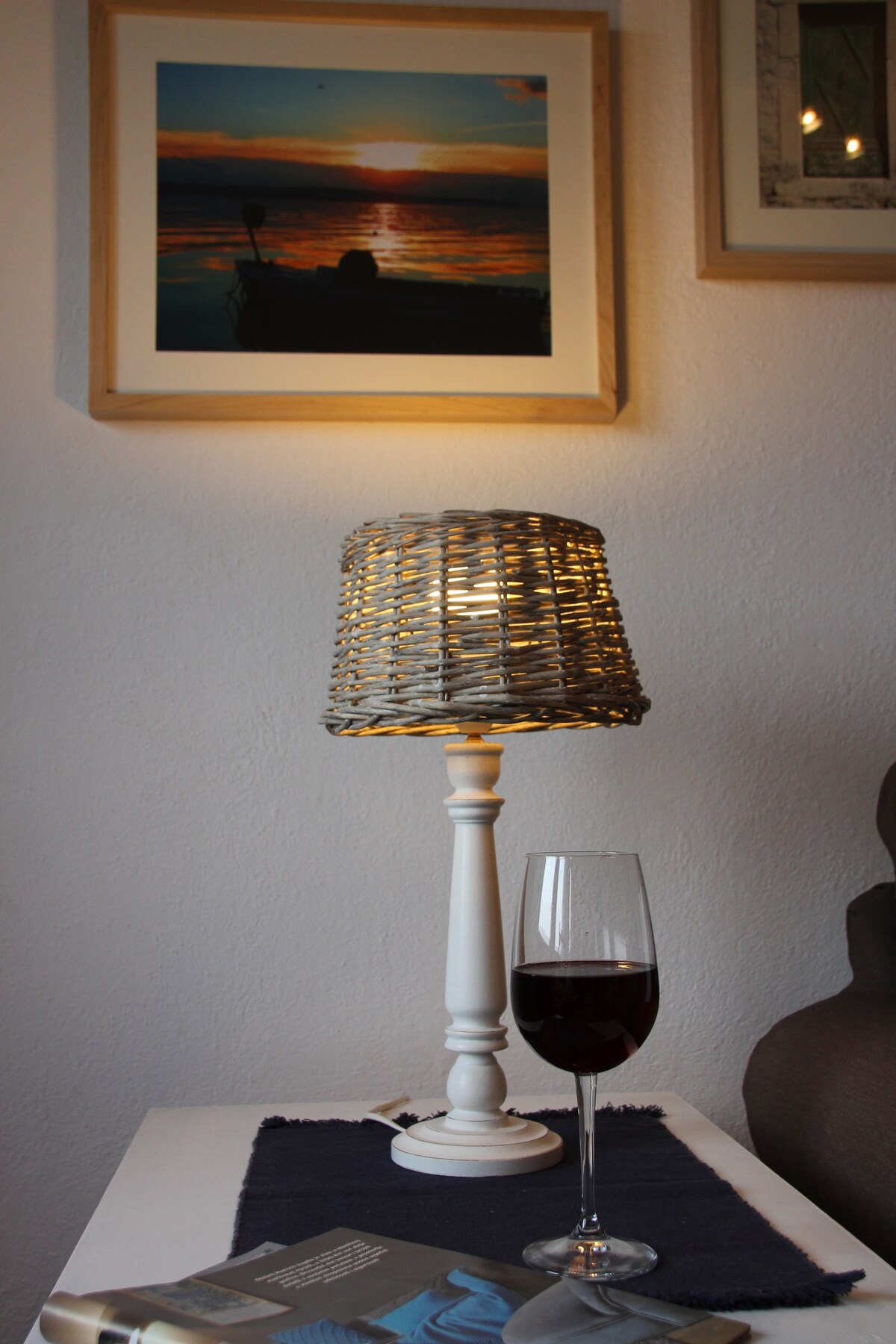 A bedside table is shown featuring a woven lamp with a warm glow, beside a glass of red wine. An open book lies on a dark blue cloth, and a framed photo of a sunset can be seen on the wall behind.