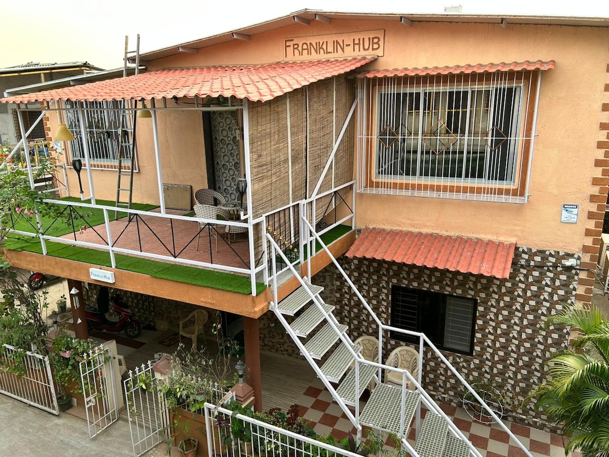 The exterior of the two-story building is showcased, featuring a textured wall design and a welcoming entrance with stairs. A covered balcony is visible, furnished with chairs, while lush greenery and a neatly maintained garden enhance the surroundings.