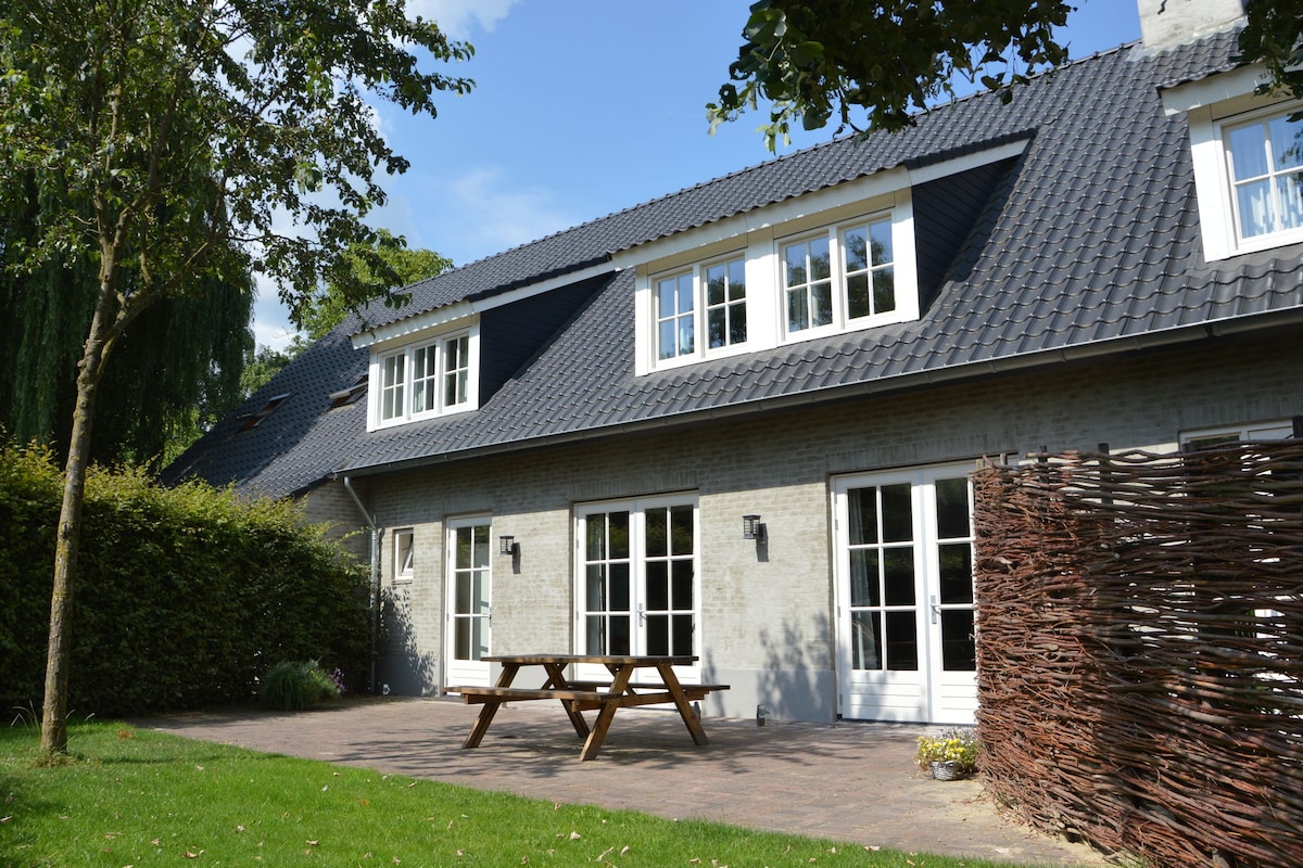 The exterior of the holiday home features a patio area with a picnic table surrounded by a well-maintained garden. Large windows allow natural light to flood the interior. A wooden fence adds privacy to the outdoor space, framed by lush green trees.
