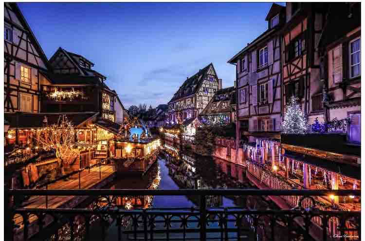 A scenic view of a charming historic village decorated for the holiday season. Twinkling lights illuminate the wooden buildings along the canals, reflecting in the water. Festive stalls display decorations, adding to the inviting atmosphere of this serene evening setting.