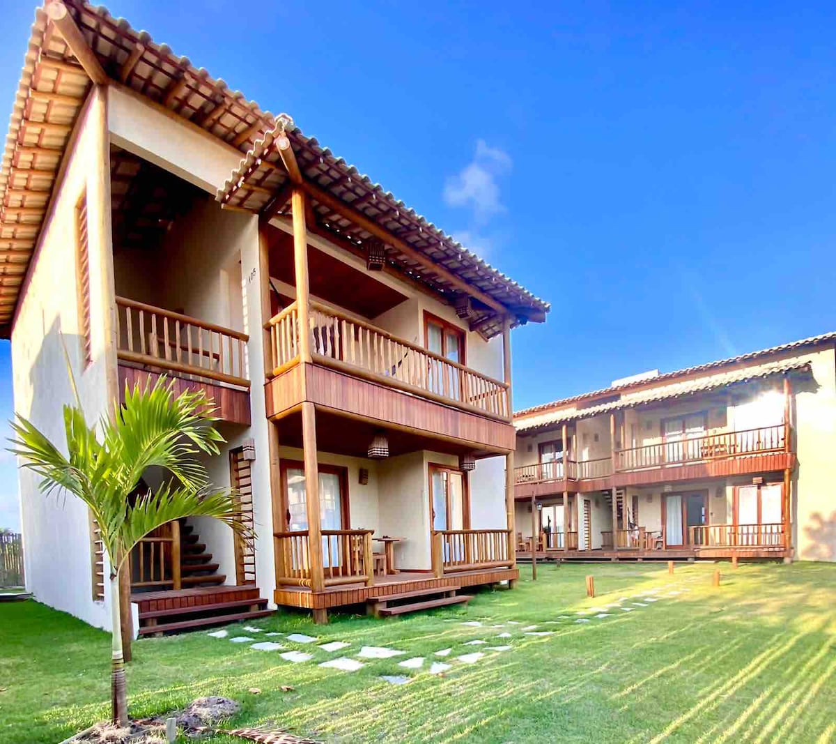 A two-story building features wooden balconies and a sloped tiled roof, surrounded by well-maintained grass. Pathways made of stone lead to the entrance. Nearby, an additional structure with a similar design complements the tranquil setting.